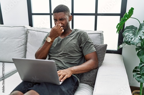 Young african american man using laptop at home sitting on the sofa smelling something stinky and disgusting, intolerable smell, holding breath with fingers on nose. bad smell