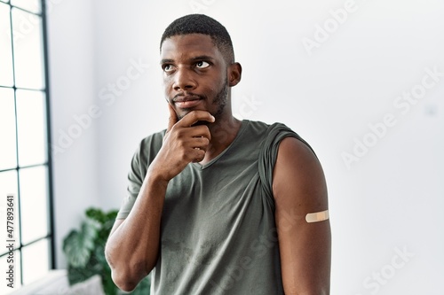 Young african american man getting vaccine showing arm with band aid serious face thinking about question with hand on chin, thoughtful about confusing idea