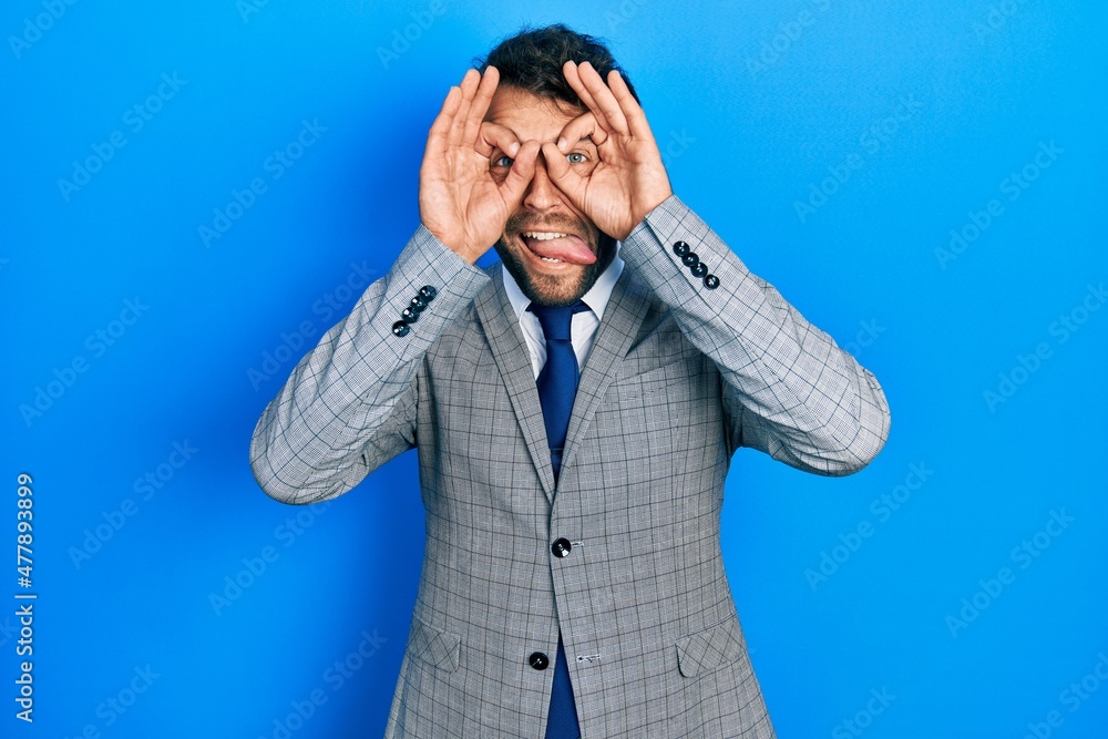 Handsome man with beard wearing business suit and tie doing ok gesture like binoculars sticking tongue out, eyes looking through fingers. crazy expression. Handsome man with beard wearing business suit and tie doing ok gesture like binoculars sticking tongue out, eyes looking through fingers. crazy expression.