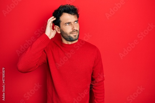 Handsome man with beard wearing casual red sweater confuse and wondering about question. uncertain with doubt, thinking with hand on head. pensive concept. © Krakenimages.com