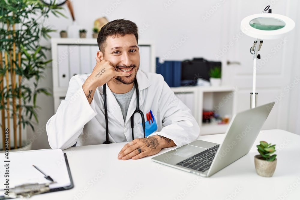 Young doctor working at the clinic using computer laptop smiling doing phone gesture with hand and fingers like talking on the telephone. communicating concepts. Young doctor working at the clinic using computer laptop smiling doing phone gesture with hand and fingers like talking on the telephone. communicating concepts.
