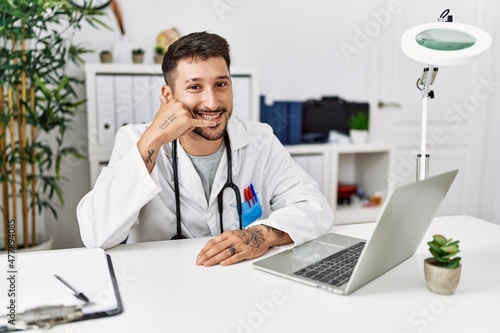 Young doctor working at the clinic using computer laptop smiling doing phone gesture with hand and fingers like talking on the telephone. communicating concepts.