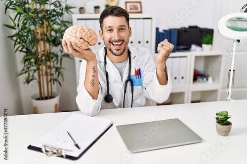 Young doctor holding brain at medical clinic screaming proud, celebrating victory and success very excited with raised arms