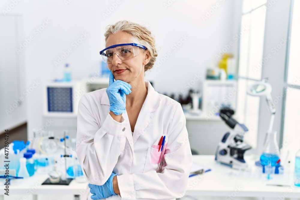 Middle age blonde woman working at scientist laboratory with hand on chin thinking about question, pensive expression. smiling with thoughtful face. doubt concept. Middle age blonde woman working at scientist laboratory with hand on chin thinking about question, pensive expression. smiling with thoughtful face. doubt concept.