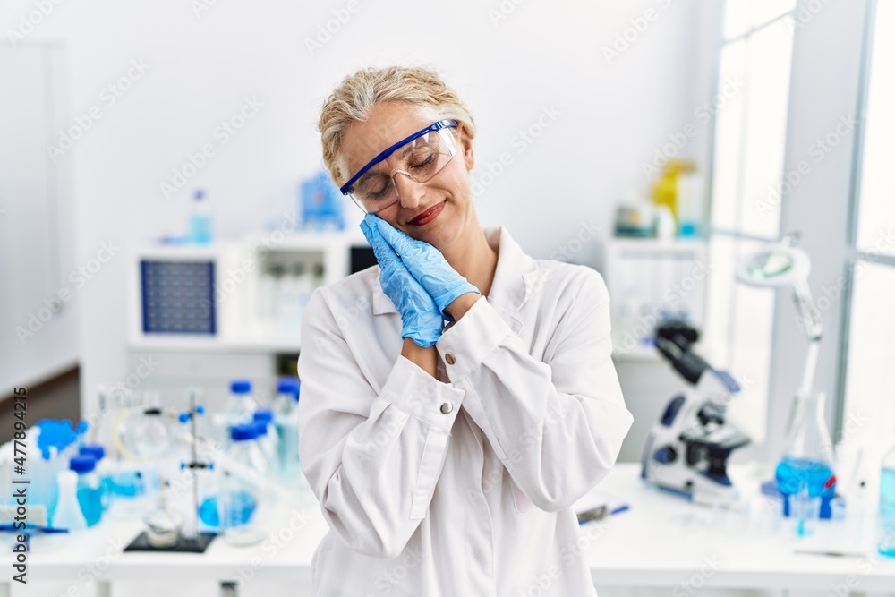 Middle age blonde woman working at scientist laboratory sleeping tired dreaming and posing with hands together while smiling with closed eyes. Middle age blonde woman working at scientist laboratory sleeping tired dreaming and posing with hands together while smiling with closed eyes.