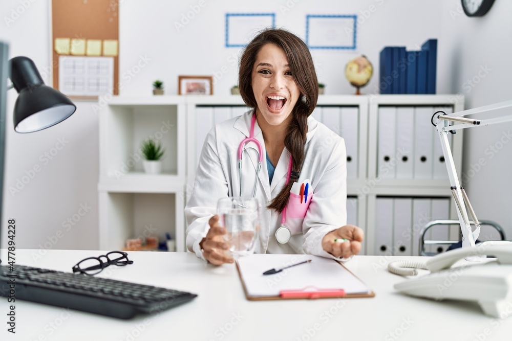 Young doctor woman holding glass of water and prescription pills celebrating crazy and amazed for success with open eyes screaming excited. Young doctor woman holding glass of water and prescription pills celebrating crazy and amazed for success with open eyes screaming excited.