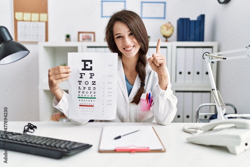 Young doctor woman holding eyesight test smiling with an idea or question pointing finger with happy face, number one Young doctor woman holding eyesight test smiling with an idea or question pointing finger with happy face, number one