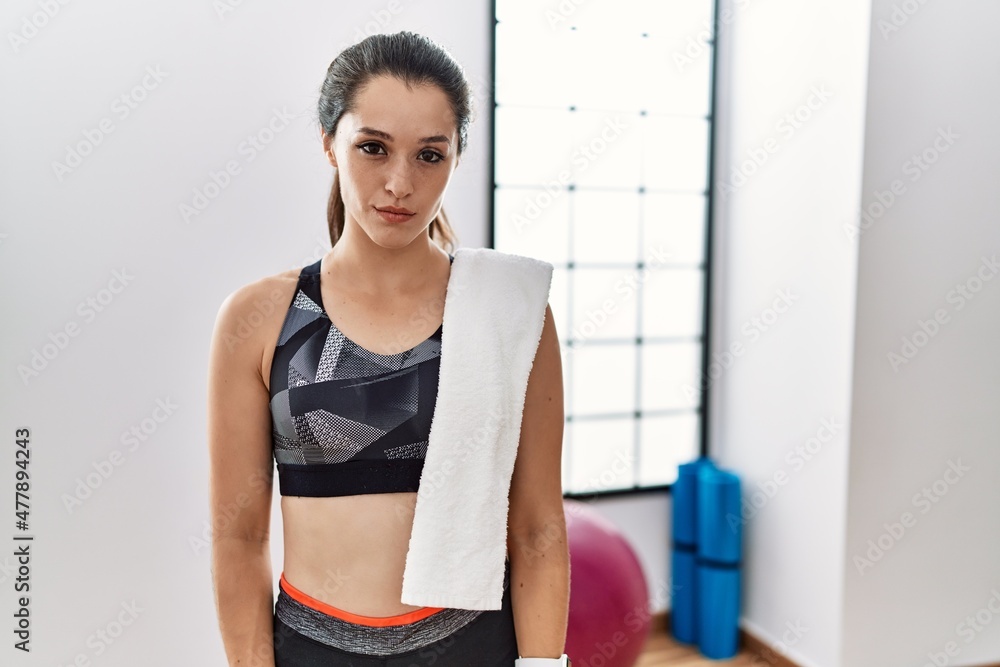 Young brunette woman wearing sportswear and towel at the gym relaxed with serious expression on face. simple and natural looking at the camera.