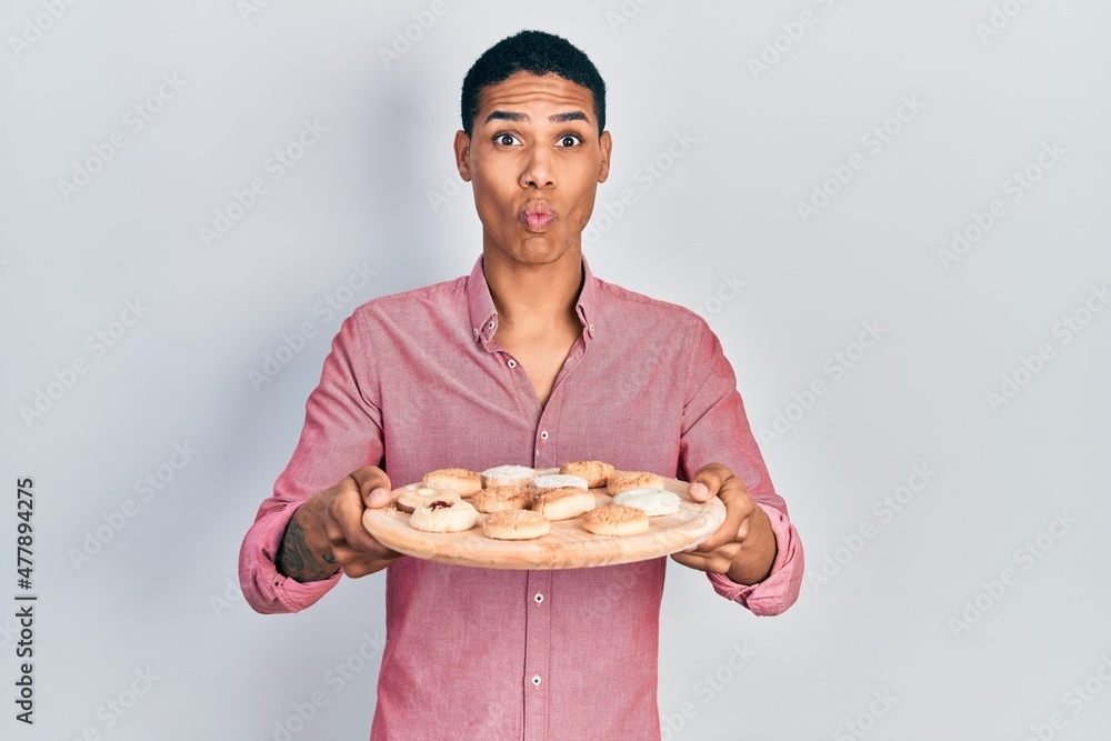 Young african american guy holding tray with cake sweets making fish face with mouth and squinting eyes, crazy and comical. Young african american guy holding tray with cake sweets making fish face with mouth and squinting eyes, crazy and comical.