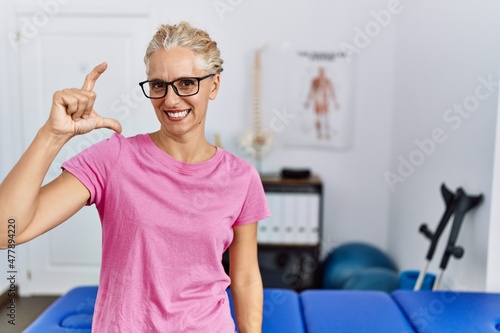 Middle age blonde woman at pain recovery clinic smiling and confident gesturing with hand doing small size sign with fingers looking and the camera. measure concept.