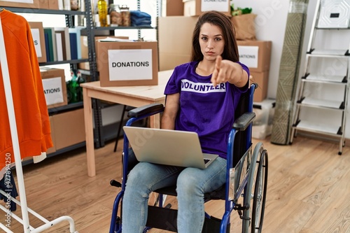 Young brunette woman as volunteer on donations stand sitting on wheelchair pointing with finger to the camera and to you, confident gesture looking serious