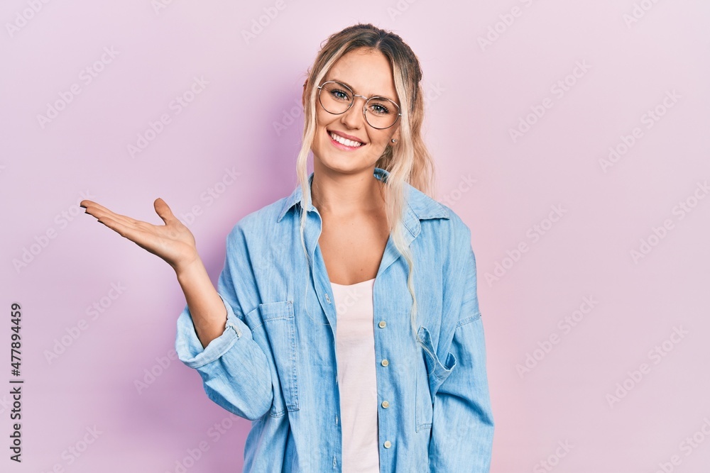 Beautiful young blonde woman wearing casual clothes and glasses smiling cheerful presenting and pointing with palm of hand looking at the camera. Beautiful young blonde woman wearing casual clothes and glasses smiling cheerful presenting and pointing with palm of hand looking at the camera.