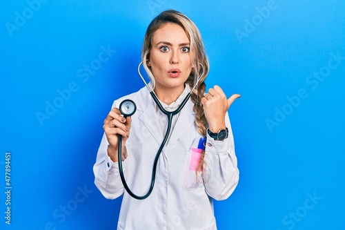 Beautiful young blonde doctor woman holding stethoscope surprised pointing with hand finger to the side  open mouth amazed expression.