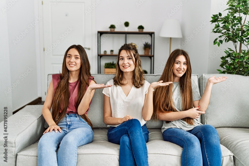 Group of three hispanic girls sitting on the sofa at home smiling cheerful presenting and pointing with palm of hand looking at the camera. Group of three hispanic girls sitting on the sofa at home smiling cheerful presenting and pointing with palm of hand looking at the camera.