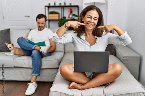 Hispanic middle age couple at home, woman using laptop smiling cheerful showing and pointing with fingers teeth and mouth. dental health concept.