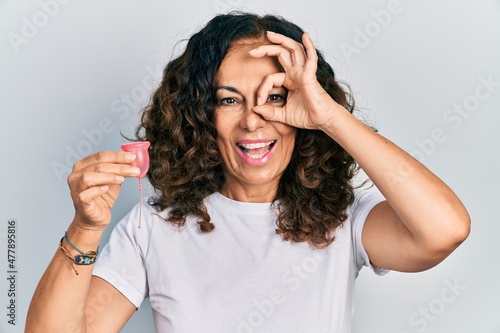 Middle age hispanic woman holding menstrual cup smiling happy doing ok sign with hand on eye looking through fingers photo