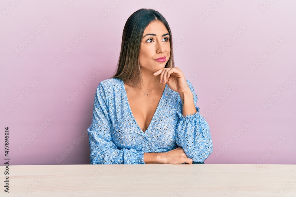 Young hispanic woman wearing casual clothes sitting on the table serious face thinking about question with hand on chin, thoughtful about confusing idea Young hispanic woman wearing casual clothes sitting on the table serious face thinking about question with hand on chin, thoughtful about confusing idea