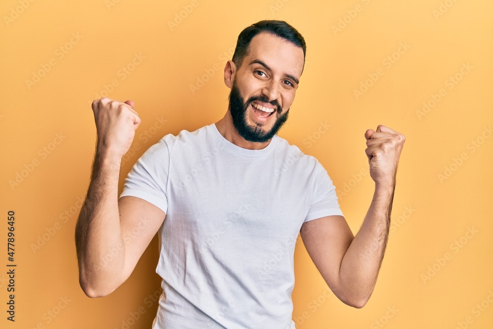 Young man with beard wearing casual white t shirt screaming proud, celebrating victory and success very excited with raised arms Young man with beard wearing casual white t shirt screaming proud, celebrating victory and success very excited with raised arms