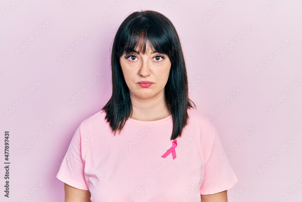 Fototapeta premium Young hispanic woman wearing pink cancer ribbon on shirt thinking attitude and sober expression looking self confident