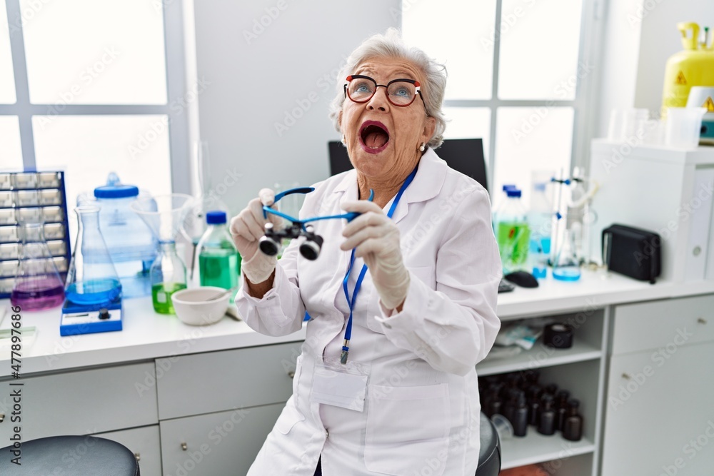 Senior woman with grey hair working at scientist laboratory using magnifying glasses angry and mad screaming frustrated and furious, shouting with anger looking up. Senior woman with grey hair working at scientist laboratory using magnifying glasses angry and mad screaming frustrated and furious, shouting with anger looking up.