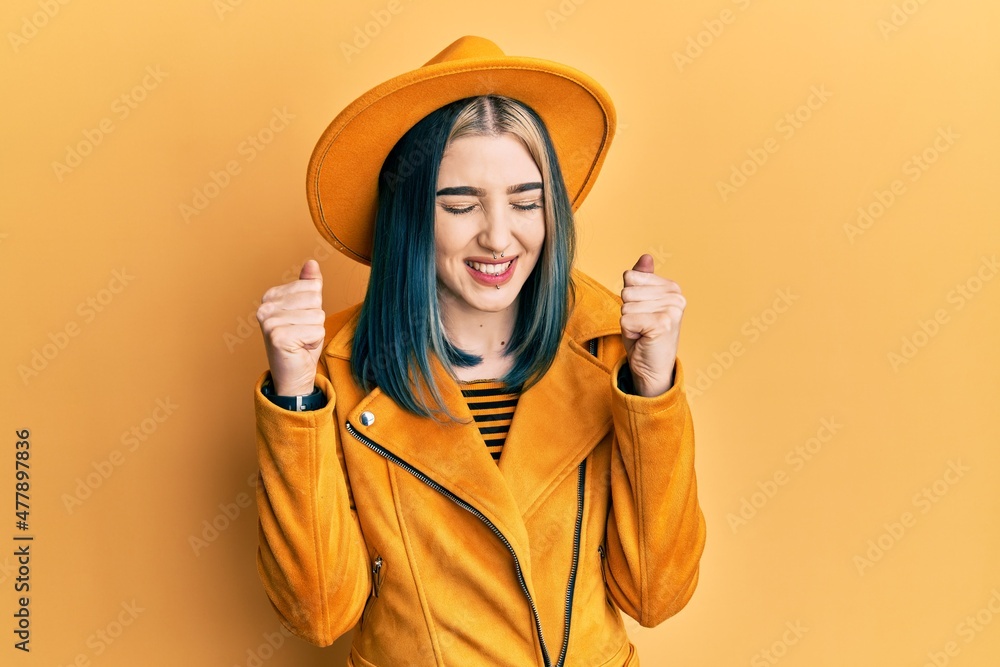 Young modern girl wearing yellow hat and leather jacket excited for success with arms raised and eyes closed celebrating victory smiling. winner concept. Young modern girl wearing yellow hat and leather jacket excited for success with arms raised and eyes closed celebrating victory smiling. winner concept.