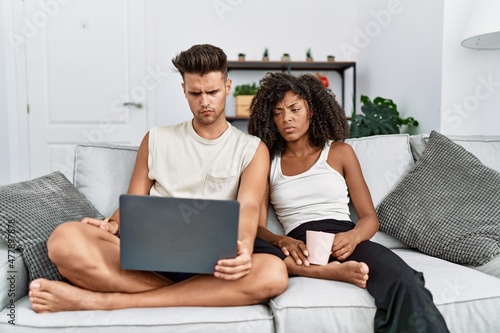 Young interracial couple using laptop at home sitting on the sofa skeptic and nervous, frowning upset because of problem. negative person.