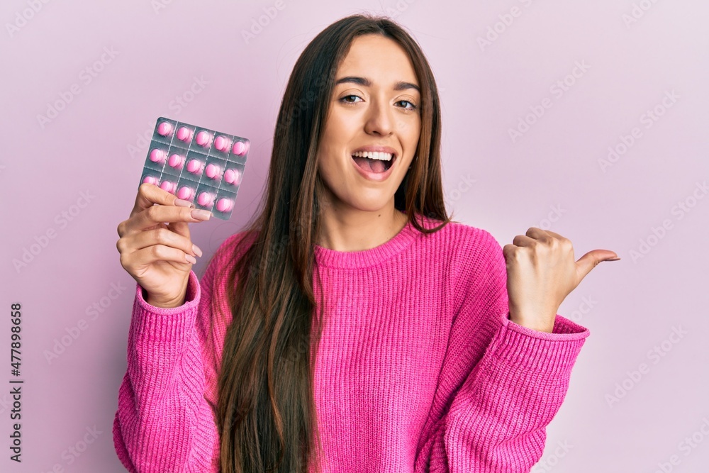 Young hispanic girl holding pills pointing thumb up to the side smiling happy with open mouth Young hispanic girl holding pills pointing thumb up to the side smiling happy with open mouth