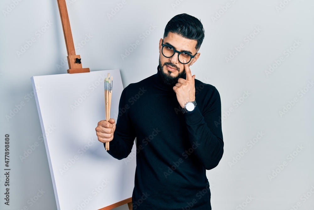 Handsome man with beard holding brushes close to easel stand pointing to the eye watching you gesture, suspicious expression Handsome man with beard holding brushes close to easel stand pointing to the eye watching you gesture, suspicious expression