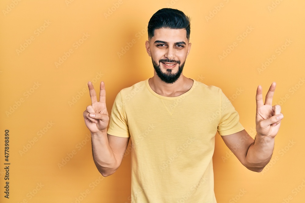 Handsome man with beard wearing casual yellow t shirt smiling looking to the camera showing fingers doing victory sign. number two. Handsome man with beard wearing casual yellow t shirt smiling looking to the camera showing fingers doing victory sign. number two.