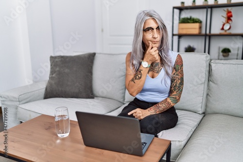 Middle age grey-haired woman using laptop at home looking confident at the camera smiling with crossed arms and hand raised on chin. thinking positive.