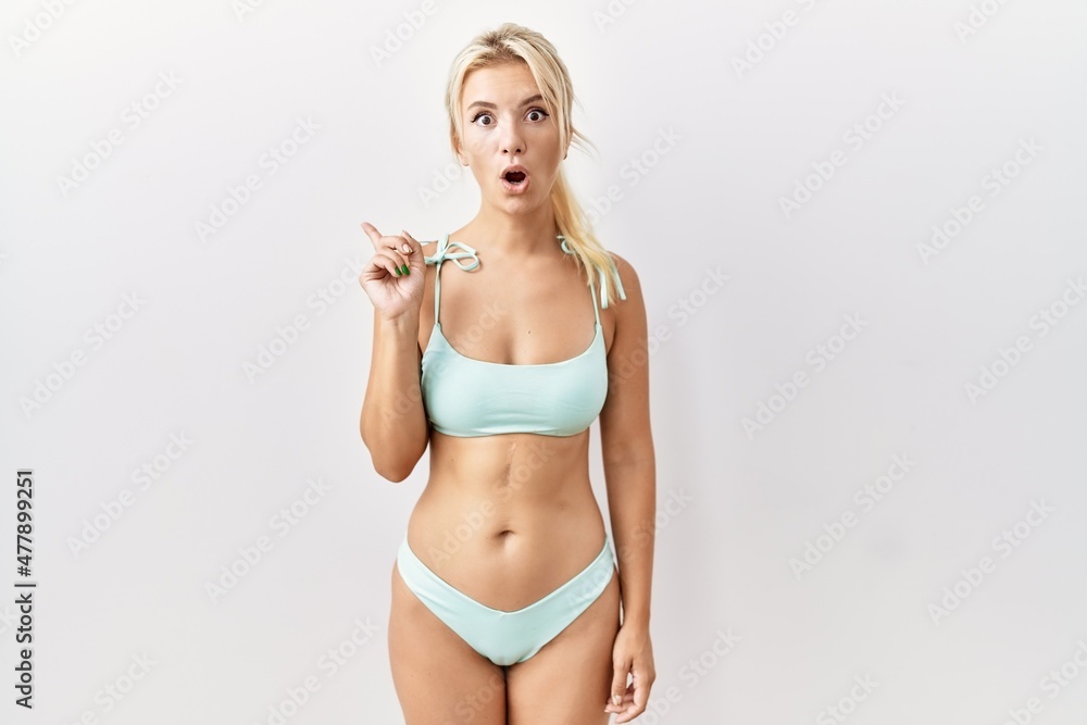Naklejka premium Young caucasian woman wearing bikini over isolated background surprised pointing with finger to the side, open mouth amazed expression.