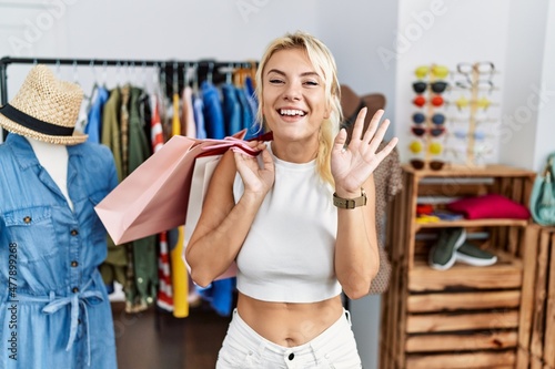 Young caucasian woman holding shopping bags at retail shop waiving saying hello happy and smiling, friendly welcome gesture