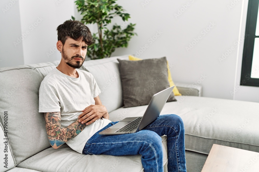 Hispanic man with beard sitting on the sofa with hand on stomach because nausea, painful disease feeling unwell. ache concept.