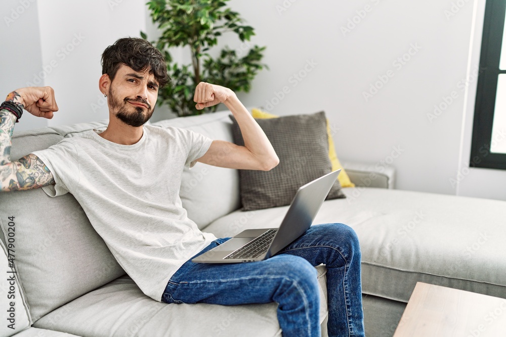 Hispanic man with beard sitting on the sofa showing arms muscles smiling proud. fitness concept. Hispanic man with beard sitting on the sofa showing arms muscles smiling proud. fitness concept.