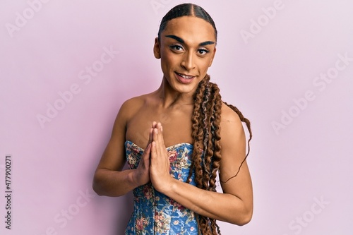 Hispanic man wearing make up and long hair wearing elegant corset praying with hands together asking for forgiveness smiling confident.