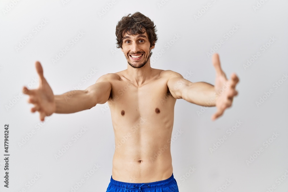 Fototapeta premium Young hispanic man standing shirtless over isolated, background looking at the camera smiling with open arms for hug. cheerful expression embracing happiness.