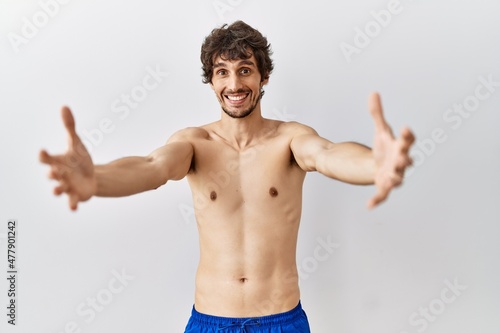 Young hispanic man standing shirtless over isolated, background looking at the camera smiling with open arms for hug. cheerful expression embracing happiness.
