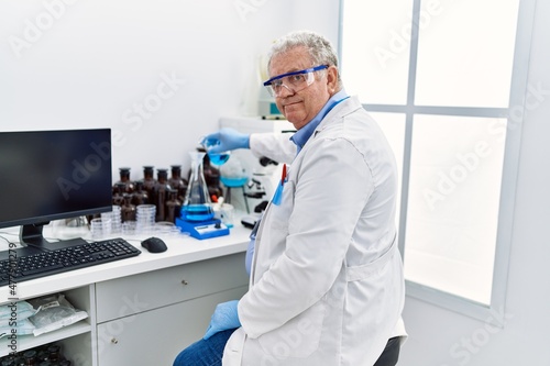 Middle age grey-haired man wearing scientist uniform pouring liquid on test tube at laboratory