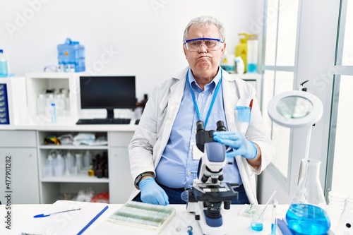 Senior caucasian man working at scientist laboratory puffing cheeks with funny face. mouth inflated with air, crazy expression.