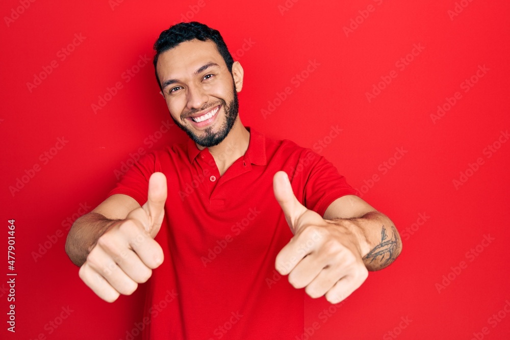 Hispanic man with beard wearing casual red t shirt approving doing positive gesture with hand, thumbs up smiling and happy for success. winner gesture. Hispanic man with beard wearing casual red t shirt approving doing positive gesture with hand, thumbs up smiling and happy for success. winner gesture.