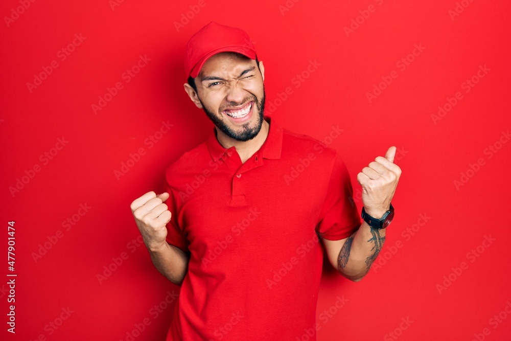 Hispanic man with beard wearing delivery uniform and cap very happy and excited doing winner gesture with arms raised, smiling and screaming for success. celebration concept. Hispanic man with beard wearing delivery uniform and cap very happy and excited doing winner gesture with arms raised, smiling and screaming for success. celebration concept.