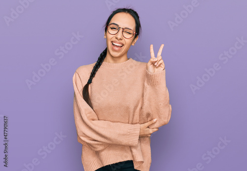 Young hispanic girl wearing casual clothes and glasses smiling with happy face winking at the camera doing victory sign. number two.