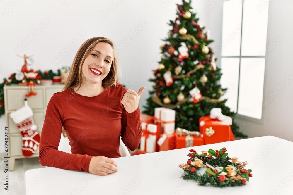Caucasian young blonde woman sitting on the table by christmas tree doing happy thumbs up gesture with hand. approving expression looking at the camera showing success. Caucasian young blonde woman sitting on the table by christmas tree doing happy thumbs up gesture with hand. approving expression looking at the camera showing success.