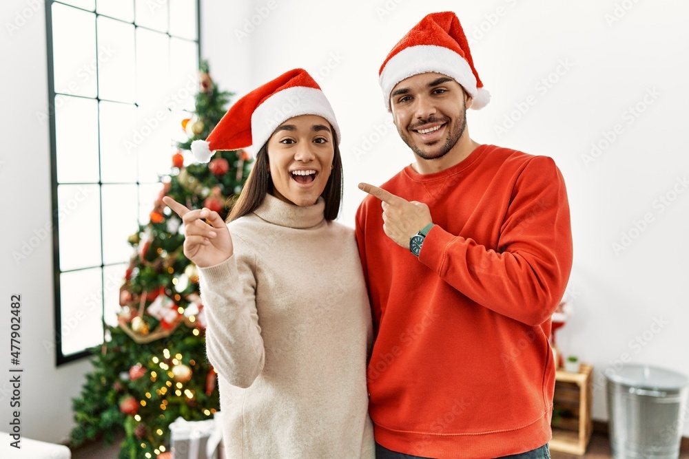 Young hispanic couple standing by christmas tree with a big smile on face, pointing with hand finger to the side looking at the camera. Young hispanic couple standing by christmas tree with a big smile on face, pointing with hand finger to the side looking at the camera.
