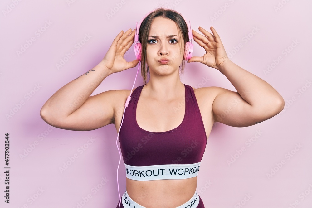 Young hispanic girl wearing gym clothes and using headphones puffing cheeks with funny face. mouth inflated with air, catching air. Young hispanic girl wearing gym clothes and using headphones puffing cheeks with funny face. mouth inflated with air, catching air.