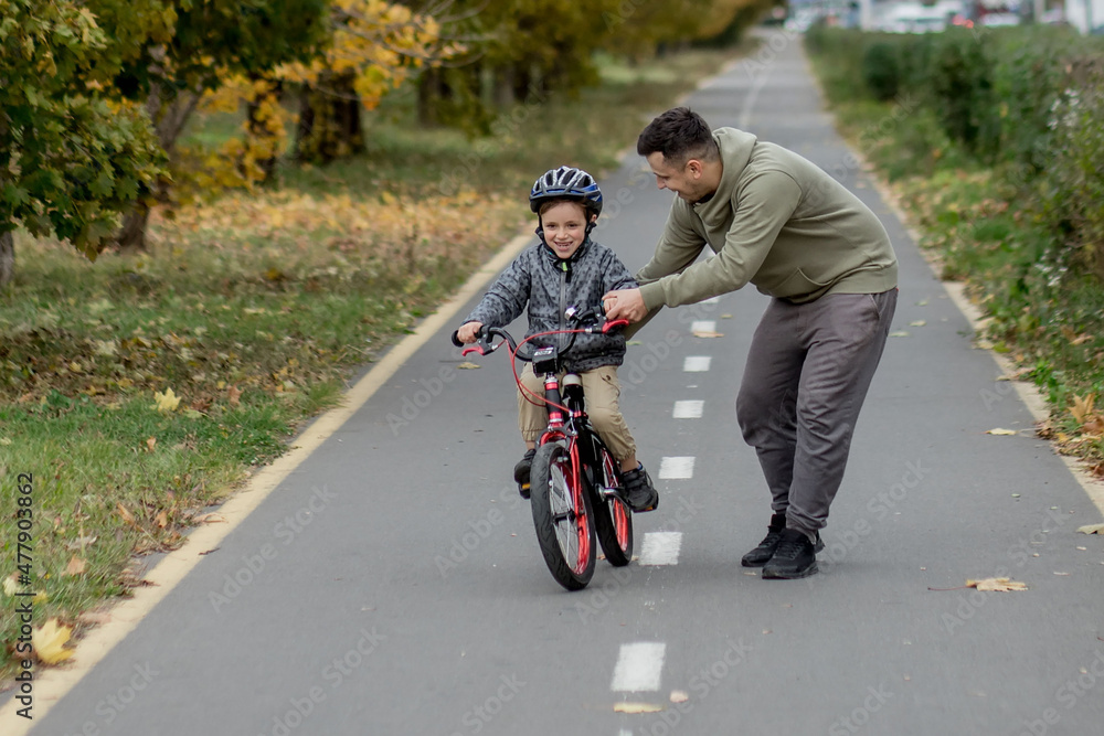 Fototapeta premium Father teaches his son to ride a bicycle on the bike path in the park. The father is holding a bicycle and the son is sitting on it