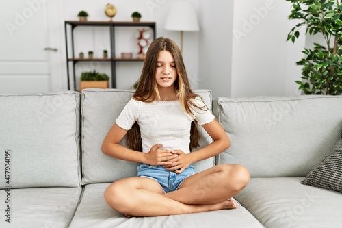 Young brunette teenager sitting on the sofa at home with hand on stomach because nausea, painful disease feeling unwell. ache concept.