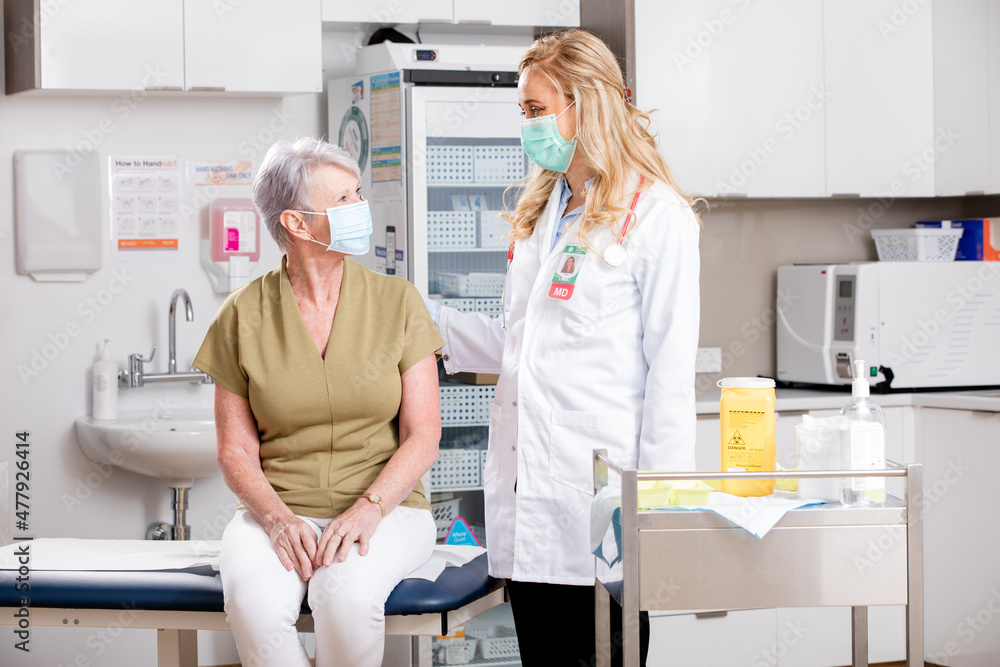 Obraz premium A Young White Female Medical Doctor Consults with an Elderly Senior Female Patient about Covid-19 Vaccine Wearing Generic ID Badge, Lab Coat, Gloves and Mask in Hospital or Health Clinic.