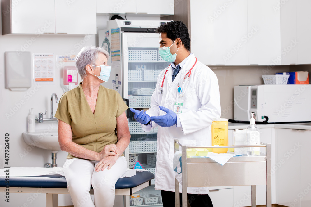 Obraz premium A Young Indian Male Medical Doctor Consults with an Elderly Senior Female Patient about Covid-19 Vaccine Injection Wearing Generic ID Badge, Lab Coat, Gloves and Mask in Hospital or Health Clinic.