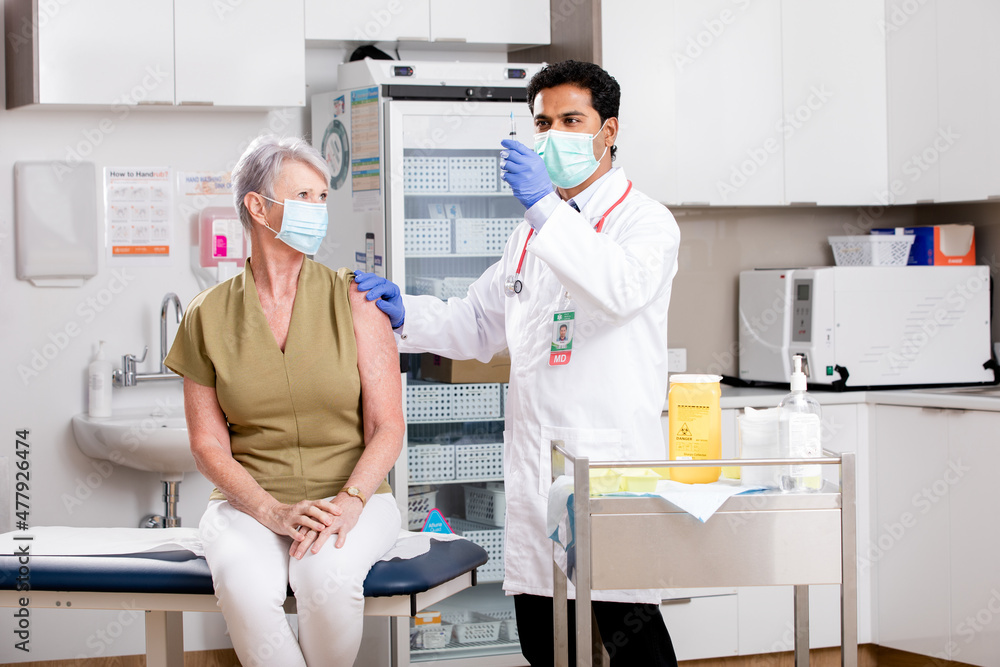 Obraz premium A Young Indian Male Medical Doctor Administering a Covid-19 Vaccine with a Syringe Needle to an Elderly Senior Female Patient Wearing Generic ID Badge, Gloves and Mask in Hospital or Health Clinic.